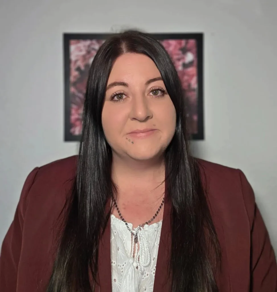 Kim Bitter mit langen dunklen Haaren im weinroten Blazer, vor heller Wand mit gerahmtem Blumenbild.