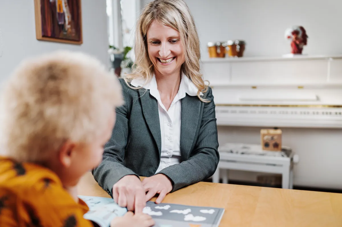 Ramona Steiner sitzt lächelnd einem Kind am Tisch gegenüber und zeigt auf Bildkarten, im Hintergrund ein Klavier im Therapieraum.