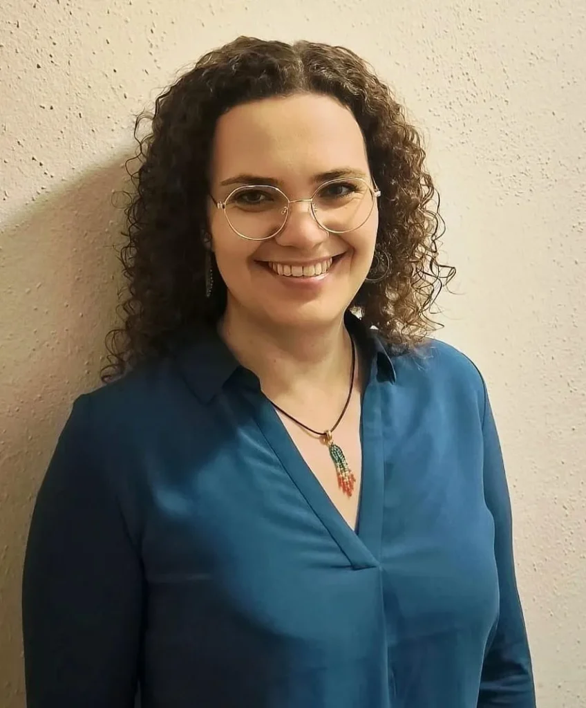 Kathrin Manhartseder mit lockigen Haaren und Brille, lächelnd in petrolfarbener Bluse vor heller Wand.